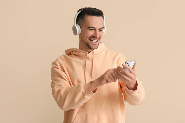 Ευτυχισμένος νεαρός άνδρας με ακουστικά χρησιμοποιώντας το κινητό τηλέφωνο σε μπεζ φόντο - Φωτογραφία, εικόνα