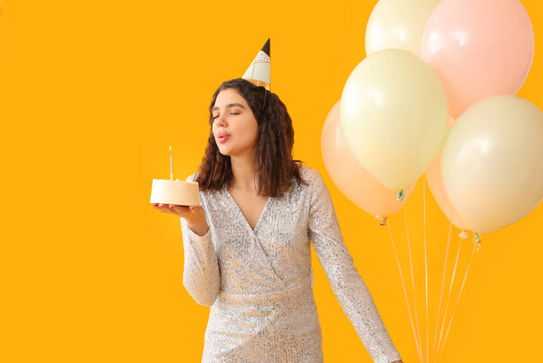 Belle jeune femme en chapeau de fête avec gâteau sucré et ballons d'air célébrant l'anniversaire sur fond jaune - Photo, image