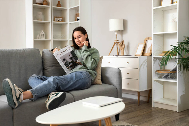 Mujer joven leyendo revista en sofá gris en la sala de estar - Foto, Imagen