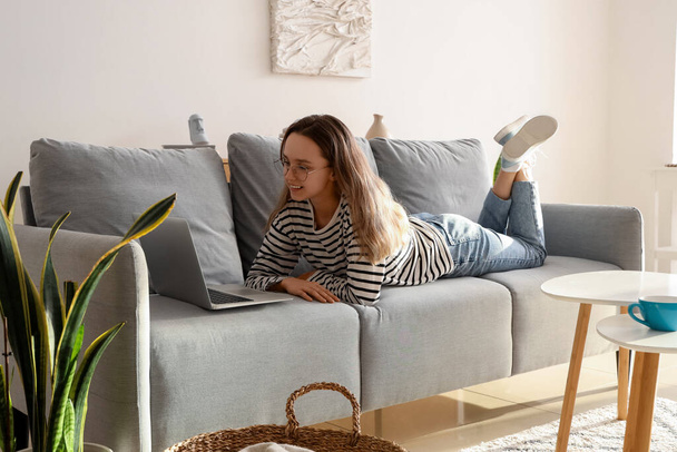 Nuori nainen silmälaseissa käyttäen kannettavaa tietokonetta harmaalla sohvalla kotona - Valokuva, kuva