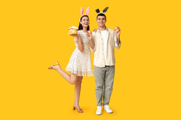 Joyeux jeune couple avec des oreilles de lapin de Pâques, des œufs et du gâteau sur fond jaune - Photo, image