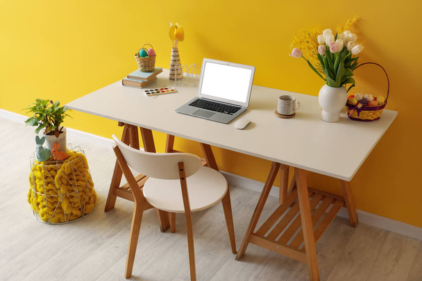 Milieu de travail avec ordinateur portable, oeufs de Pâques et fleurs près du mur jaune dans le bureau - Photo, image