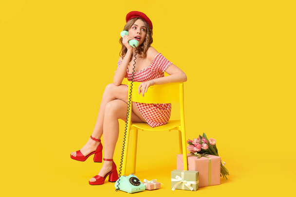 Young woman talking by retro telephone with gift boxes and bouquet of beautiful tulips on chair against yellow background. International Women's Day - Photo, Image