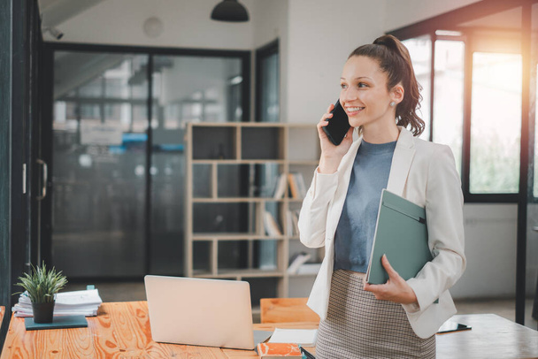 Lächelnde junge Geschäftsfrau in einem Telefonat, während sie einen Ordner in der Hand hält und neben ihrem Laptop in einer Büroumgebung steht - Foto, Bild