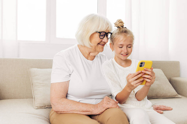 Бабушка селфи вместе космос ребенок телефон видео-звонок футболка с фотографией диван связи белый внучка обнимая в помещении семейное образование улыбаясь копия - Фото, изображение
