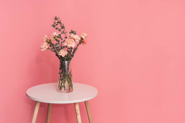 Букет гвоздик с эвкалиптовой веткой в стеклянной вазе на круглом столе на розовом фоне. - Фото, изображение