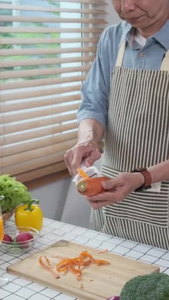 ásia sênior homem no avental peeling alguns cenouras para fazer cenoura suco no cozinha. - Filmagem, Vídeo