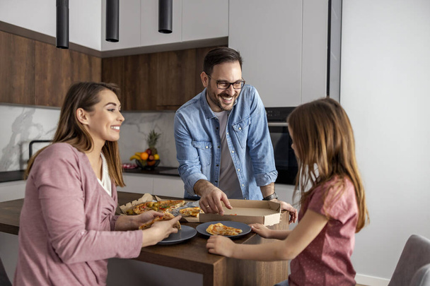 Mama, tata i córka jedzą razem w domu. Szczęśliwa rodzina je pizzę w domu. - Zdjęcie, obraz