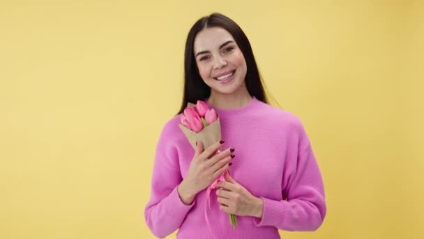 Adorabile signora bruna in rosa indossare sorridente e tenendo bouquet di tulipani mentre in piedi in studio. Donna sentirsi amata e bella mentre si gode i fiori dal fidanzato amato su sfondo giallo. - Filmati, video