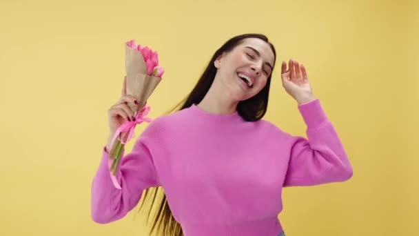 Radostná fena držící krásnou tulipánovou kytici a tančící štěstím. Potěšená žena čichající květiny a vyjadřující spokojenost s přítomností při stání ve studiu přes žluté pozadí. - Záběry, video