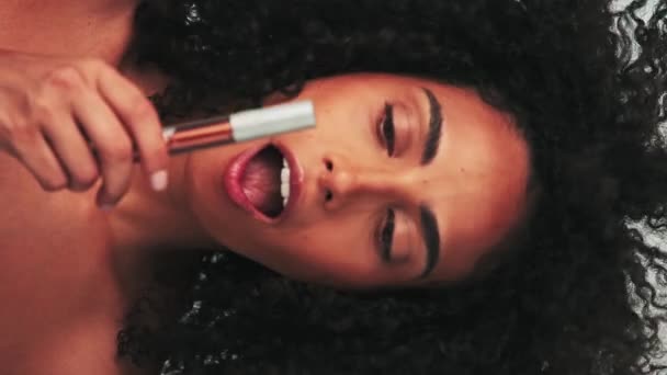 Retrato vertical de una joven modelo con pelo afro lúgubre y maquillaje brillante. Pintura cubos con lápiz labial. Moción lenta, primer plano. Captura de estudio. - Metraje, vídeo