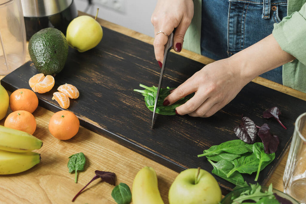 Mujer cortando hojas de espinaca verde, preparando batido vegano saludable con espinacas, plátano, manzana y aguacate en una licuadora sobre una mesa. - Foto, Imagen