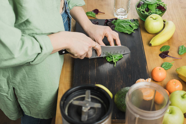 Mujer cortando hojas de espinaca verde, preparando batido vegano saludable con espinacas, plátano, manzana y aguacate en una licuadora sobre una mesa. - Foto, imagen