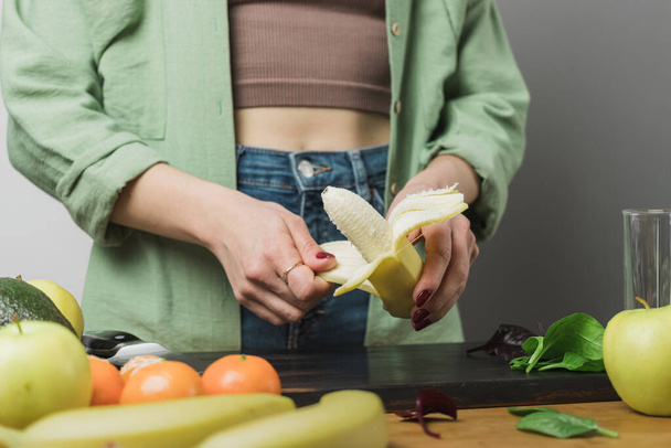 Mujer pelando plátano, preparando batido vegano saludable con hojas de espinaca, manzana, mandarina y aguacate en una mesa. - Foto, imagen