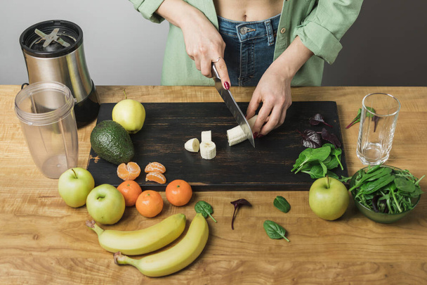 Mulher cortando banana, preparando smoothie vegan saudável com folhas de espinafre, maçã e abacate em uma mesa. - Foto, Imagem