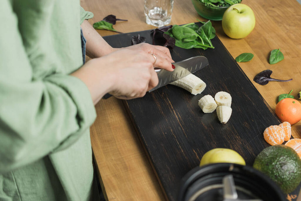 Женщина режет банан, готовит здоровый веганский смузи со шпинатом листья, яблоко и авокадо на столе. - Фото, изображение