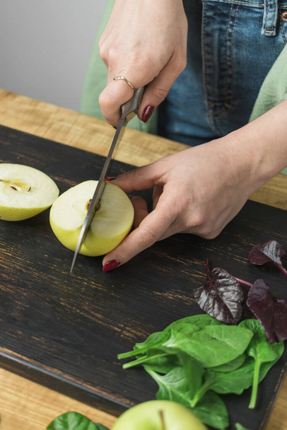 Woman cutting apple, preparing healthy vegan smoothie with spinach leaves on a table. - Photo, Image