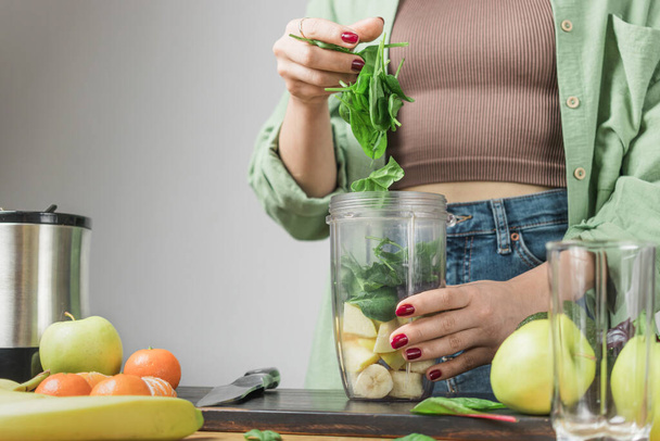 Vrouw met groene spinazie bladeren, het bereiden van gezonde veganistische smoothie met spinazie, banaan, appel en avocado in een blender op een tafel. - Foto, afbeelding