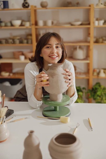 Retrato de una joven alfarera en delantal con una taza mirando a la cámara mientras posa en el taller - Foto, imagen