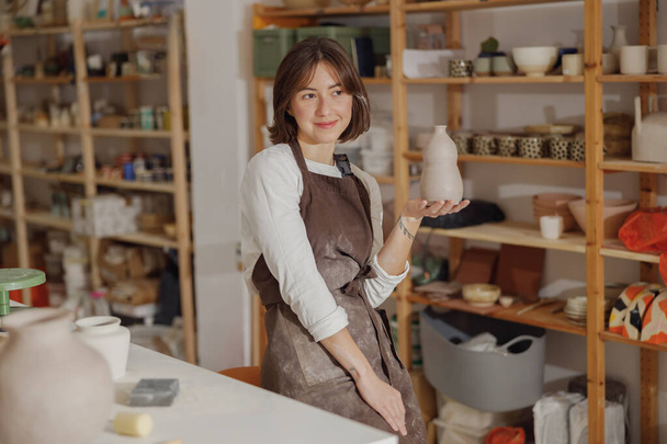 Улыбающийся предприниматель ремесел женщина держит кружку в керамической студии, а смотрит в сторону  - Фото, изображение