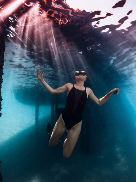 Freediver kobieta pozowanie pod wodą pod molo w błękitnym oceanie z promieni słonecznych. - Zdjęcie, obraz
