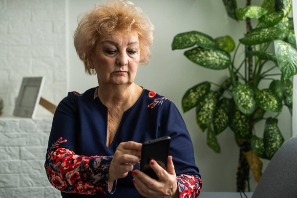 Χαλαρή ώριμη γυναίκα 60 ετών, ηλικιωμένη μεσήλικη πελάτισσα που κρατάει smartphone χρησιμοποιώντας εφαρμογή για κινητά, γραπτό μήνυμα, προσφορές ηλεκτρονικού εμπορίου αναζήτησης σε συσκευή τεχνολογίας κινητών τηλεφώνων που κάθεται στον καναπέ στο σπίτι. - Φωτογραφία, εικόνα