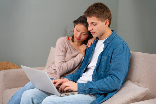 Young multiracial couple sits on the couch at home using a laptop surfing the wireless internet on a gadget - Photo, Image