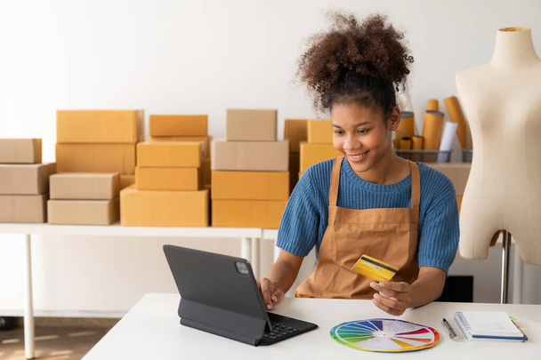 Young African woman running an online store, startup, small business, SME, using a smartphone or tablet, works with receipt boxes and checks online orders to prepare to pack boxes to sell to customer - Photo, Image