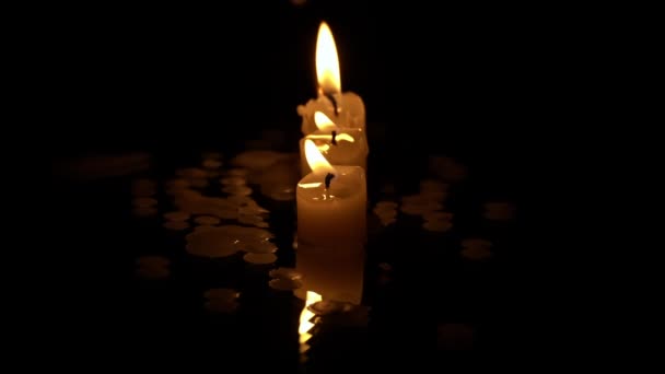 Drie paraffinekaarsen branden op rij op een zwarte achtergrond met reflectie. Drie witte kaarsen branden met een feloranje vlam op een donkere achtergrond. Symbool van verdriet, herinnering aan de doden, oorlogsconcept - Video