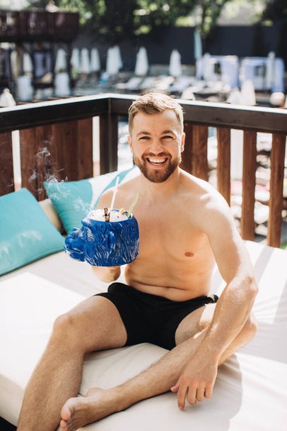 Όμορφος νεαρός που χαλαρώνει με ένα κοκτέιλ δίπλα στην πισίνα στις διακοπές. - Φωτογραφία, εικόνα