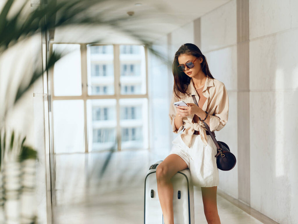 Esperando: Joven viajera sentada en la terminal del aeropuerto con su equipaje y usando un smartphone, esperando su vuelo. - Foto, imagen