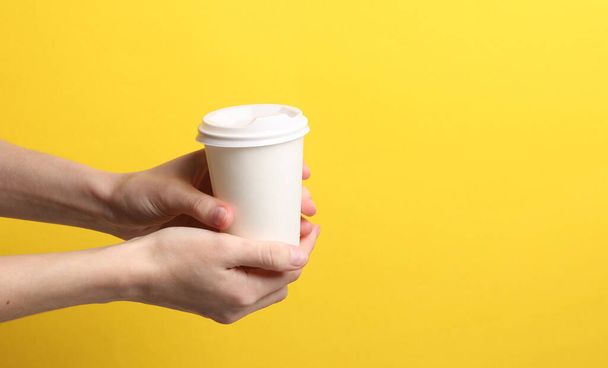 Mano sosteniendo blanco Desechable sacar taza para bebidas calientes maqueta (té, café) sobre fondo amarillo - Foto, imagen