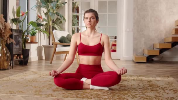 Une femme en tenue de sport rouge fait une méditation matinale pour organiser ses pensées et augmenter sa concentration avant le début de la journée de travail. Une jeune femme assise sur un tapis en position lotus - Séquence, vidéo