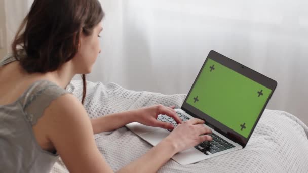 緑の画面のラップトップに入力する若い女性の背面図。若いブルネットの女性はベッドの上に横たわっている間、ラップトップでリモートで動作します。高品質のフルHD映像 - 映像、動画