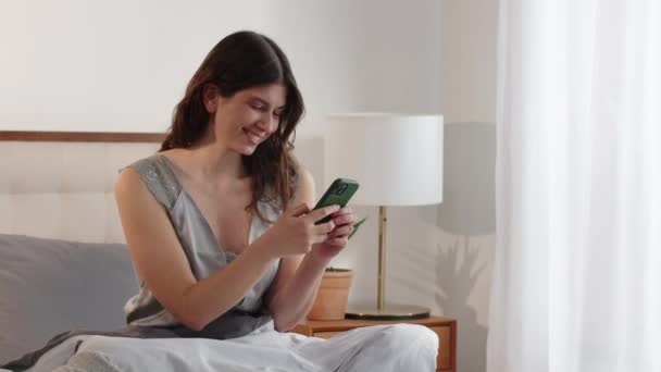 Une belle femme souriante est assise sur le lit et parcourt les médias sociaux sur son smartphone. Une brune tapant un message téléphonique avec le sourire. Des images FullHD de haute qualité - Séquence, vidéo