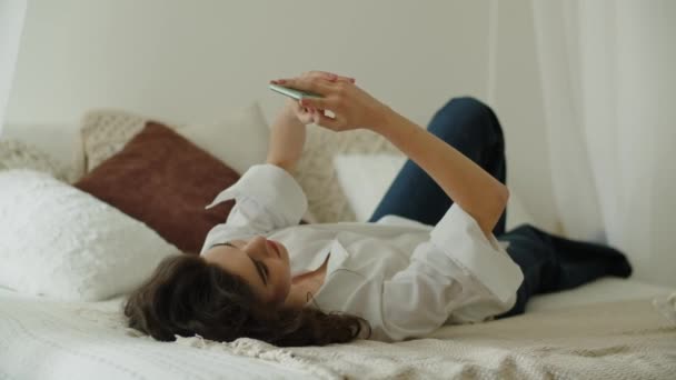 Charmantes Mädchen lächelt, während es mit jemandem im Schlafzimmer SMS schreibt. Frau mit Smartphone im Bett. Hochwertiges 4k Filmmaterial - Filmmaterial, Video
