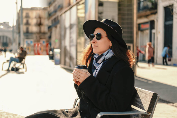 Calm young woman wearing hat and coat enjoys her morning coffee while sitting in the street. - Photo, Image