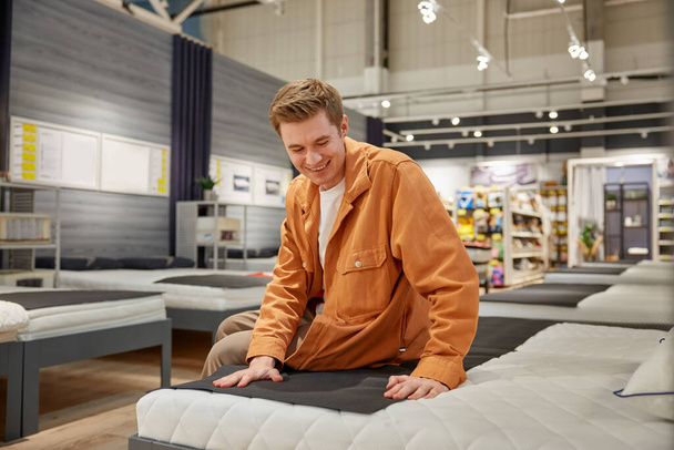Handsome male customer sitting on new bed at furniture store. Young man buyer choosing orthopedic mattress for bedstead to buy - Photo, Image