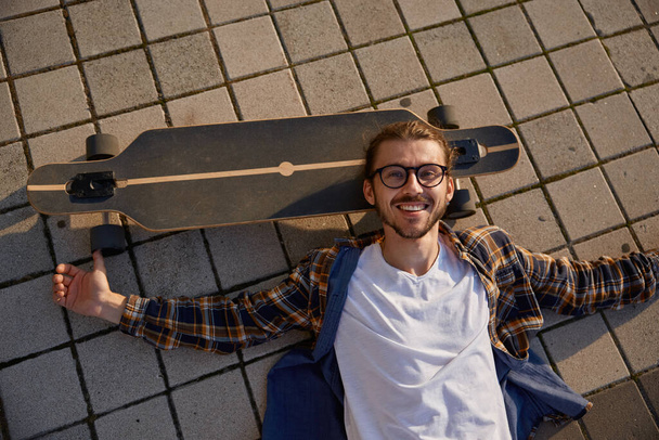 Tired hipster guy lying on skateboard after training relaxing in sunset high angle view. Skateboarding hobby and leisure time of young people - Photo, Image