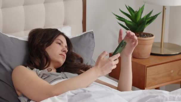 Nuori nainen tarttuu puhelimeen ja sammuttaa sen hälyttimen. Herännyt nainen makaa sängyssä ja katsoo ulos ikkunasta. Laadukas FullHD kuvamateriaalia - Materiaali, video