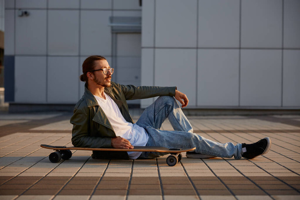 Le hipster à la mode se repose avec le skateboard assis sur la route asphaltée profitant d'une soirée d'été calme après l'entraînement de skateboard. Concept de culture urbaine et de style jeunesse - Photo, image
