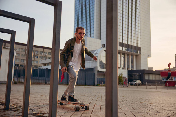 Cool τύπος μετακινούμενος στο skateboard πάνω από το σύγχρονο υπόβαθρο της πόλης. Νεαρός όμορφος άνδρας οδήγηση longboard διατήρηση της ισορροπίας κατά τη διάρκεια της ταχύτητας - Φωτογραφία, εικόνα