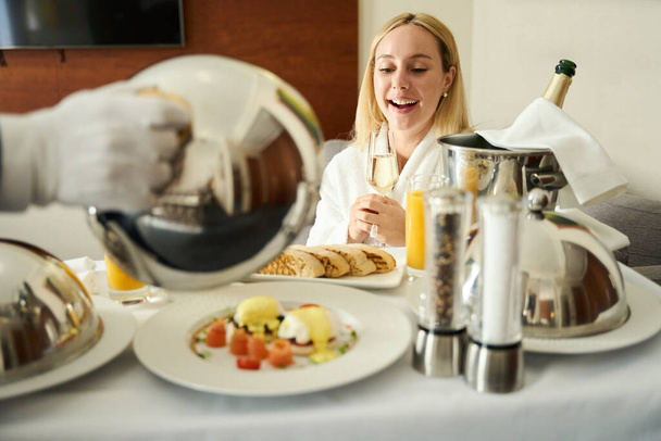Σερβίροντας πρωινό με σαμπάνια στο δωμάτιο σε ένα τραπέζι σερβιρίσματος, ο επισκέπτης του ξενοδοχείου βρίσκεται στο χώρο χαλάρωσης - Φωτογραφία, εικόνα