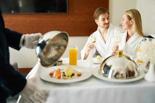 Kellner serviert den Frischvermählten in ihrem Zimmer auf einem mobilen Tisch das Frühstück, ein Mann und eine Frau genießen Champagner - Foto, Bild