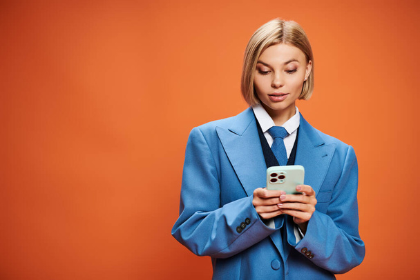 positive junge Frau mit blonden kurzen Haaren posiert mit ihrem Smartphone auf orangefarbenem Hintergrund - Foto, Bild