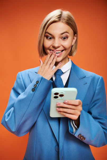 pozytywne dobrze ubrana kobieta z krótkimi blond włosy w eleganckim stroju trzyma telefon na pomarańczowym tle - Zdjęcie, obraz