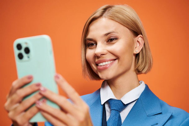 femme joliment sophistiquée avec des cheveux blonds courts en tenue chic tenant le téléphone sur fond orange - Photo, image