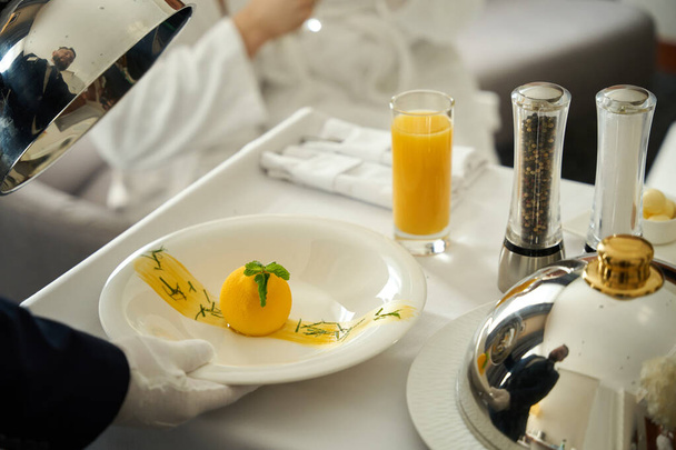 Σερβιτόρος σερβίρει πρωινό σε έναν επισκέπτη του ξενοδοχείου στο κρεβάτι, έναν άνδρα που φοράει λευκά γάντια - Φωτογραφία, εικόνα