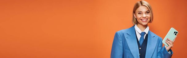 mujer elegante feliz con pelo corto y rubio en traje elegante sosteniendo el teléfono sobre fondo naranja, pancarta - Foto, imagen