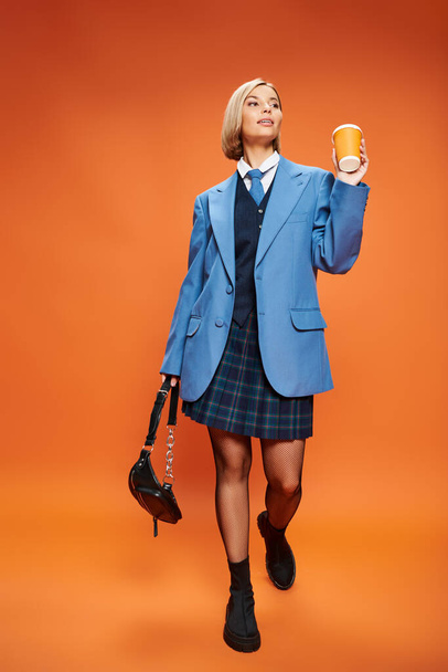 radostná sofistikovaná žena s blond krátké vlasy drží kabelku a kávu na oranžovém pozadí - Fotografie, Obrázek